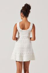 A woman is wearing a sleeveless, white dress with a fitted waist and a flared skirt. The dress features intricate lace and embroidered patterns throughout, with a scalloped hemline and wide, scalloped shoulder straps.